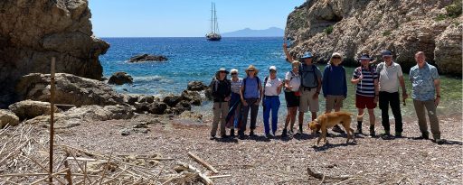 A group of guests and a dog standing on a pebbled beach between rocky cliffs, after doing a beach clean. There is a gulet on the blue sea behind them.