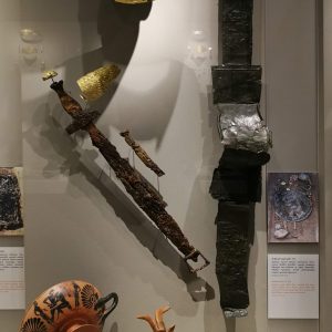 The full display of (male) Grave 131, with helmet, gold sheet covering the face, gold wrist-cover, iron sword and dagger, bronze handle of a composite shield, clay figurine of a ram and an Attic black-figure kylix (wine cup) depicting Theseus slaying the Minotaur. Circa 530 BC.