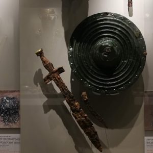 The full display of (male) Grave 145, with helmet, gold sheet covering the face, iron sword, dagger and spearhead, bronze shield, bronze 'ritual vessel', local clay drinking cup, Corinthian alabastra (perfume flasks) and Athenian black-figure wine cup. Circa 550-530 BC.