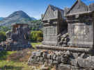 Ancient stone tombs with gabled roofs stand prominently in the foreground of the ruins of Sidyma. Behind them, a rocky mountain peak towers over a sun-drenched valley filled with greenery and the remains of other weathered stone structures.