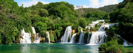 Wide shot of the Krka waterfalls, with green water in the foreground and green trees behind, near Sibenik in Croatia