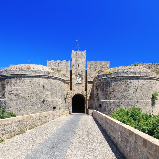 A head on view of a fortified gateway of medieval Rhodes, viewed from over a stone bridge. All is of brownish grey stone, neatly cut and jointed. Most prominent are the two squat drum towers either side of the entrance, looking very thick and impregnable. There are largely blank – very minimal openings. They are capped, almost literally, by low domed roofs, looking like lids to barrels thanks to the distinct band of stone where they meet the drums proper. The gate itself is dark and tall with a rounded arch. Over it stands a tall, narrow square tower, rising higher than the drums; it is flanked by broader square towers a storey shorter. All have swallow-tail merlons and are pierced by thin arrow slits. Just above the gate is an ogival plaque in paler stone, where the relief of an angel can barely me made out. Two dark vertical slits flank it, the former channels for the drawbridge mechanism. Once again the sky is a deep, cloudless blue.