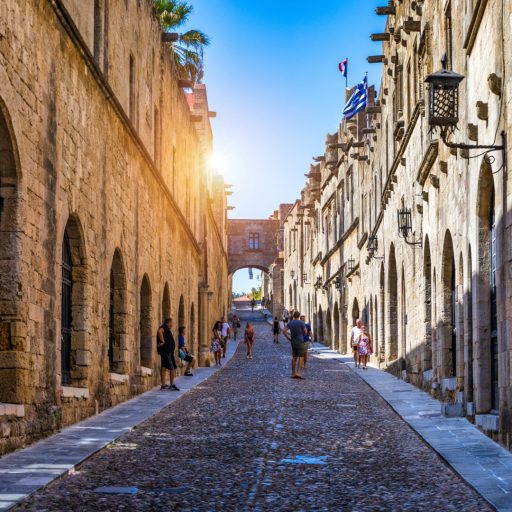 A mediaeval honey-coloured stone street of medieval Rhodes, rising away from our viewpoint. It is all of stone, rising vertically for two storeys either side but pierced with dark, pointed-arch entryways, particularly uniform on the left. The sky is a cloudless azure with a blazing sun directly ahead. The storeys are marked with a moulding course, and the upper storey has oblong windows. Black iron openwork lanterns project over the street from brackets set at intervals over the doors on the right. The street itself is cobbled but flanked with slabbed pavements. About halfway up on the right, one building has small turrets on its upper storey. From this one flutter the flags of Greece and France.