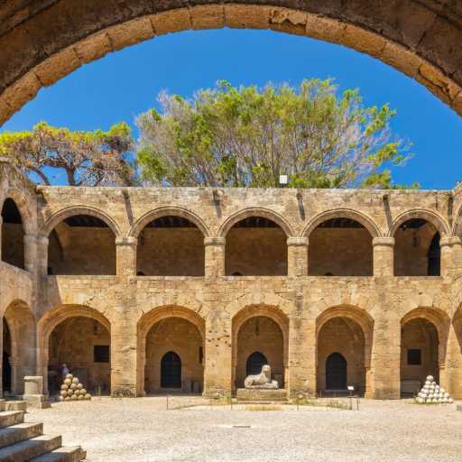 An open courtyard in medieval Rhodes; over it is an intensely blue sky, framed by an arch over our heads. The material is honey-coloured stone, set off by a spray of green tree branches above it. There are two storeys of arcading framing it: we are looking straight on at one of these. The upper arcade has squatter arches, but with decorated mouldings springing from cushion-like capitals. The lower arcade shades a walkway pierced by dark pointed-arch doorways. The courtyard itself is bleached by the sun; at the corner are stacks of cannonballs in paler stone. At the centre, before a doorway and facing us directly is the recumbent stone statue of a lion, its head turned to face us directly.
