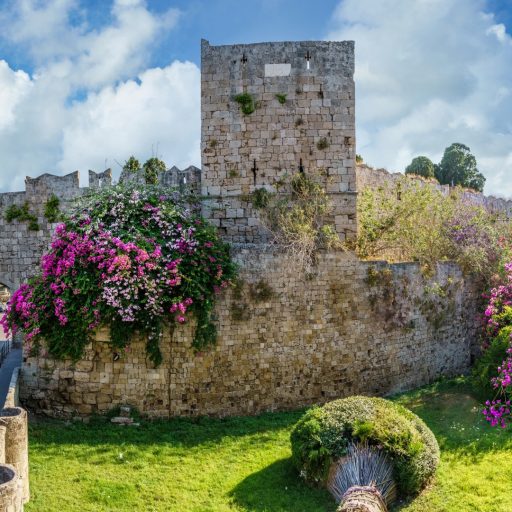 A tall medieval angular, square tower rising up above the corner of medieval Rhodes’ fortifications. It’s pierced with two arrow slits on its middle and upper levels. It’s clearly been rebuilt several times – the whole structure is an overall grey brown, but the individual stones very through a granite grey, beige and dark brown with no particular pattern and deeply-etched joins; several sections have wildly different coursing patterns where considerable rebuilding has taken place. The tower is flanked by battlements with zig-zagged tops like a swallow’s tail. It stands over a grassy flat-bottomed ditch and by a bridge to the left; the sky is blue with white, fluffy clouds. Over the fortifications flow.