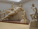 A large, dramatic ancient Roman marble sculptural group from the Sperlonga sculptures in a bright museum gallery with white walls and a skylight. The central figure is the massive, muscular cyclops Polyphemus, lounging backward in a drunken stupor across a rocky outcrop. Several smaller statues of Greek companions, led by Odysseus, surround him, dynamically working together to hoist and aim a long, thick wooden stake toward the cyclops's closed eye.