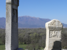 Two ancient Roman stone steles standing outdoors at the Philippi archaeological site in Macedonia, Greece. The foreground stele features a relief carving of a rider and Latin inscriptions, while the distant snow-capped Mount Pangaion rises behind them under a clear blue sky.