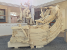 An eye-level shot of the Ship of Odysseus and Scylla from the Sperlonga sculptures. The central focus is the fragmented prow of a stone ship, upon which several weathered, dynamic human figures are positioned. One figure towards the rear of the ship leans forward intently, while another figure appears to be falling backward off the vessel amid a chaotic scene. Ancient Greek script is painted on the white wall behind the installation, and large glass windows and doorways are visible in the background.