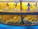 An eye-level shot of a museum display featuring a detailed cross-section model and mural of the ancient Bronze Age trading ship, Uluburun shipwreck, illustrating its cargo and crew. The top section shows a bright yellow mural of the ship's deck, where several figures in ancient attire—including crew members in white waistcloths working on the mast and sails, and elite figures in patterned robes—interact. The bottom section reveals the wooden interior of the hull packed tightly with archaeological cargo, including neatly stacked metallic ingots, large ceramic amphorae, woven baskets filled with goods, bundles of thick rope, and rows of small clay jugs.