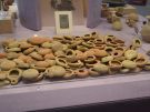 A large cluster of ancient, terracotta oil lamps is piled inside a shallow, clear acrylic display tray on a purple museum table. The lamps are small, round, and varying shades of beige, tan, and terracotta orange, with many featuring visible inventory numbers written on their surfaces. In the background, additional ancient pottery pieces, including small jugs, vases, and framed artifacts, are displayed on stands. The cult festivals of Demeter and Persephone are always accompanied by large numbers of terracotta lamps, used in the recreation of Demeter’s desperate hunt for her daughter. These are from the Central Sanctuary, still used in the Spanish period, third-first centuries BC.
