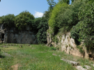 The remains of the School of Aristotle at Mieza, now grassy clearing surrounded by tall rock walls and dense green trees, with cave-like openings carved into the stone on the left and vegetation growing along the cliff face under a bright blue sky.