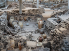 An elevated view of the prehistoric archaeological site of Akrotiri in Santorini, Greece, preserved under a large modern shelter. The scene displays excavated stone foundations and ruined walls of ancient buildings. Grouped together inside the dirt rooms are numerous large, light-colored ceramic storage jars called pithoi, some featuring subtle decorative patterns. In the background, elevated metal walkways and pillars accommodate visitors touring the covered excavation area.