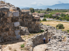 An elevated, wide shot of the ancient Cyclopean stone walls of Tiryns in Greece, looking out toward a surrounding valley. In the foreground, massive, roughly shaped gray and beige boulders form a towering fortification wall on the left. A narrow stone-lined pathway winds downward through lower stone ruins, where two visitors are walking. In the background, a flat plain features a road with driving cars, scattered modern buildings, fields, and distant blue mountains under a partially cloudy sky.