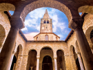 A wide-angle view of the central atrium of the Euphrasian Basilica in Poreč, featuring stone arches supported by marble columns. The 16th-century stone bell tower rises in the background against a dramatic blue and white sky.
