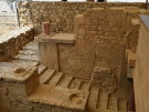 The excavated Grand Staircase at the Palace of Knossos in Crete, showing tiered stone steps, balustrades with column sockets, and dry stone walls.