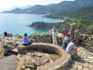 A small tour group exploring the stone ruins of Ancient Stagira, the birthplace of Aristotle, on a sunny hill overlooking the turquoise waters and forested mountains of the Halkidiki coastline in Greece.