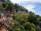 A group of hikers walking along a rocky, narrow trail on the Lycian Way in Turkey, with green Mediterranean vegetation and a view of the sea under a blue sky.