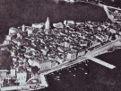 A vintage black-and-white aerial photograph of the coastal town of Poreč, Croatia, situated on a small peninsula. The image shows a dense cluster of historic stone buildings and narrow streets, with a prominent church bell tower rising above the rooflines, surrounded by the Adriatic Sea and several harbor piers.