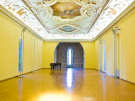 The interior of the Istrian Assembly Hall in Poreč, Croatia, featuring vibrant yellow walls, a polished wooden floor, and a black grand piano. The ornate Baroque ceiling is decorated with intricate white stuccowork and colorful frescoes in medallions, framed by arched windows.