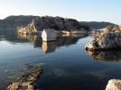 A partially submerged ancient Lycian sarcophagus in the calm, clear water of Kaleköy harbor, Turkey, with rocky islands in the background.