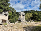Two ancient Lycian sarcophagi with distinctive crested stone lids standing in a rocky field at Kyaneai, Turkey, surrounded by trees under a cloudy sky