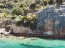 Ancient Lycian rock-cut tomb facade mimicking a wooden house, carved into a massive grey limestone cliff along the shoreline of Kekova Island, Turkey.