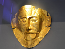 The Mask of Agamemnon, an ancient Mycenaean gold funeral mask mounted against a dark gray museum display wall. The mask is crafted from a thin sheet of hammered gold, depicting a bearded man with a prominent nose, closed almond-shaped eyes, detailed eyebrows, a stylized mustache with upturned ends, and large, detailed ears on either side. The gold surface shows minor folds and slight damage along its top edges, highlighted by a spotlight from above.