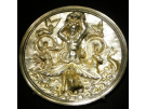 An ancient silver medallion with gilded accents against a solid black background. The intricate relief carving depicts the mythological sea monster Scylla as a female figure with her arms raised, holding an a large stone above her head. Below her waist, her body transforms into large, stylized fish tails and multiple snapping sea dogs or monsters rising from stylized, swirling waves.