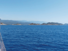 View from the bow of a Turkish gulet cruising through the deep blue Mediterranean Sea with Kekova island, smaller rocky outcrops and mountain ridges in the background under a clear sky.