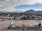 A group of people on the summit of the palace citadel of Tiryns, in Greece, centred on a square room, its walls formed of rough blocks flanked to left and right by similarly sized ones, and at the centre an irregular depression in the floor.