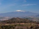The snow-capped peak of Mount Etna rising behind the stone ruins of the ancient Greek city of Morgantina in Sicily, with wind turbines visible on the distant hills.