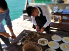 Two men on a sunny outdoor wooden deck over turquoise water are shucking fresh oysters in Ston, Croatia. One man in a white shirt and black apron carefully opens an oyster at a wooden table, where several plates with lemon wedges and shucked oysters are laid out next to a pile of unopened shells.
