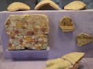 Fragments of ancient, painted wall plaster and pottery are mounted on a light purple display block inside a museum. The largest plaster fragment on the left features a dense pattern of colorful circles in shades of red, yellow, and green, resembling faux marble or stone. Two smaller fragments to the right show striped and scrolling decorative borders, while curved terracotta pieces sit along the top and in the foreground.