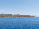 Panoramic view of a single white sailboat cruising the deep blue Mediterranean Sea between the rocky coastlines of Kekova Island and Kaleköy, Turkey.