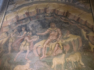An ancient Roman floor mosaic depicting a mythological scene set inside a cave with a vaulted rock ceiling. The central figure is the muscular cyclops Polyphemus, seated on a rock draped with an animal skin, possessing a prominent third eye on his forehead. To the left, three smaller figures—Odysseus and his companions—approach him, with the foremost man offering a large shallow bowl of wine. In the foreground, several sheep and goats graze along the bottom of the scene, framed by a patterned stone border.