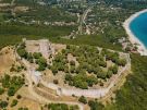 Aerial view of the historic stone crusader fortress of Platamon with a central round tower and surrounding walls on a hilltop, overlooking a long sandy beach and turquoise sea, with a coastal town, roads, and green hills stretching into the distance.