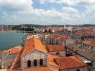 High-angle view of the historic Euphrasian Basilica in Poreč, showing its large red-tiled roof and Byzantine mosaics on the upper facade. The background features the red-tiled roofs of the Old Town, the blue Adriatic Sea, and a clear sky with white clouds.