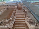 A close-up view of an ancient stone staircase at the Palace of Nestor in Pylos, Greece. The well-preserved steps lead up into the ruins of the palace's upper floors, surrounded by low stone walls and situated beneath a modern protective roof and visitor walkway.
