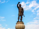 A modern bronze statue of Philip II standing on a large round pedestal, raising his right fist in triumph against a blue sky with white clouds, at Methoni in Macedonia. While besieging the city in 359 BC, he lost an eye to a defender's arrow.