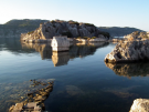 A freestanding, ancient Lycian stone sarcophagus standing partially submerged in the clear, calm shallow coastal waters near the harbor of Kaleköy, Turkey.