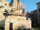 Ruins of the 1st-century Roman Temple of Neptune in Poreč, Croatia, featuring a weathered stone wall with decorative cornices under a clear blue sky, surrounded by historic buildings and grass.
