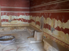 The ancient Throne Room at the Palace of Knossos in Crete, featuring a stone throne, stone benches, a large basin, and walls decorated with griffin frescoes.