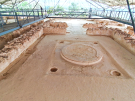 The throne room at the Mycenaean Palace of Nestor in Pylos, showcasing the well-preserved circular clay hearth and the surrounding floor plan of the megaron, with visitors observing from a raised metal walkway.