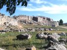 A view of the ancient citadel of Tiryns, in Greece, from lower down, strewn with boulder-like stones, and the wall rising to some considerable height, resting on a natural rise out of which craggy, scored natural rock outcrops.