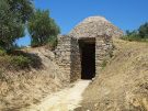 An ancient stone beehive-shaped tomb, known as a tholos tomb, with a narrow dirt pathway leading to its dark rectangular entrance. The structure is built into a grassy hillside under a clear blue sky, with a few olive trees visible on the left.