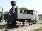 A restored 1908 narrow-gauge steam locomotive model U.37, displayed as a monument on a short track in Koper, Slovenia. The black engine features red accents on its wheels and sits against a backdrop of trees and a bright sky, representing the historic Parenzana line.