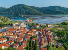 An aerial view from an ancient stone wall overlooking the town of Ston, Croatia, featuring its iconic red-tiled roofs, sprawling rectangular salt pans, and lush green hills under a clear blue sky.