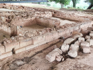 Close-up view of the excavated stone foundations at the Palace of Nestor. The image shows several small, rectangular rooms defined by low walls of light-colored stone and earth, with a modern protective metal roof structure visible at the very top.