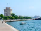 The White Tower beside Thessaloniki’s waterfront promenade, with people walking along the tree-lined path, a small boat floating in the blue sea, and city buildings and tour boats in the background under a clear sky.