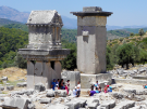 A tour group and guide at the ancient stone theatre ruins at Xanthos, Turkey, featuring the Harpy Tomb pillar and a panoramic view of the Lycian valley.